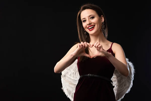 Femme souriante en robe avec des ailes montrant geste de coeur isolé sur noir — Photo de stock