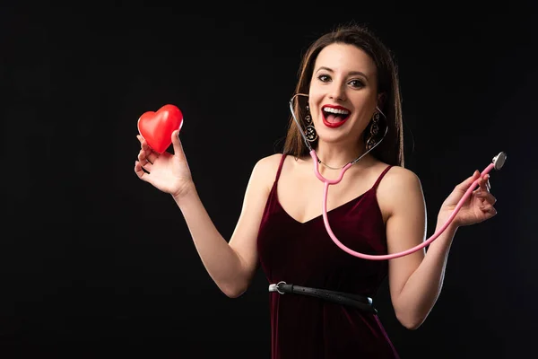 Улыбающаяся женщина в платье держит модель в форме сердца и стетоскоп изолирован на черном — стоковое фото