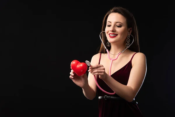 Smiling woman in dress holding heart-shaped model and stethoscope isolated on black — Stock Photo