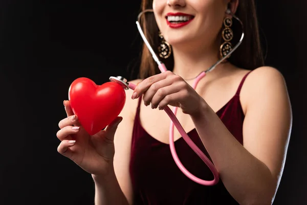 Обрезанный вид улыбающейся женщины в платье, держащей модель в форме сердца и стетоскоп, изолированный на черном — стоковое фото