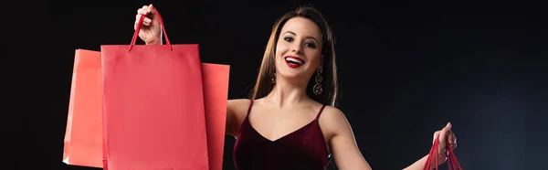 Panoramic shot of smiling woman in dress holding shopping bags on black background — Stock Photo