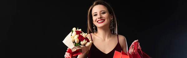 Panoramic shot of smiling woman in dress holding shopping bags and bouquet on black background — Stock Photo