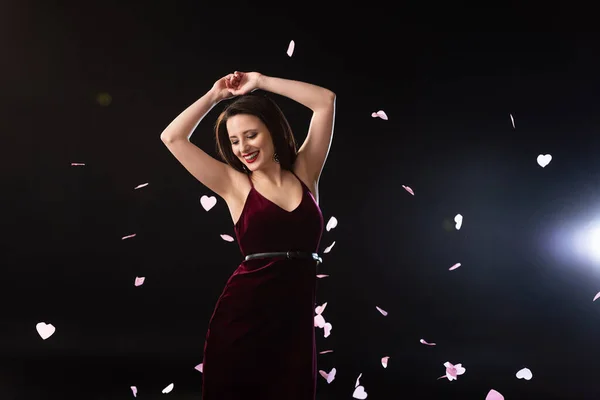 Sorrindo mulher no vestido de pé perto de cair confete no fundo preto — Fotografia de Stock