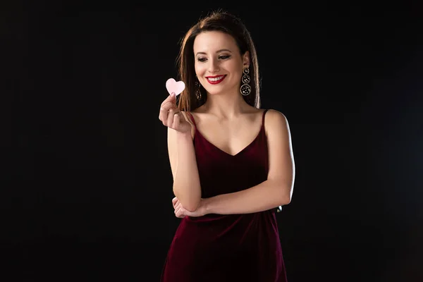 Smiling woman in dress holding heart-shaped card isolated on black — Stock Photo