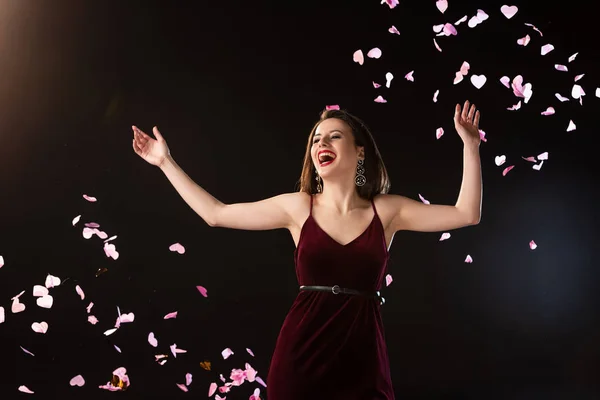 Smiling woman in dress standing near falling confetti on black background — Stock Photo