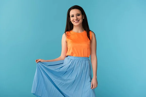 Smiling and attractive woman looking at camera and holding skirt on blue background — Stock Photo