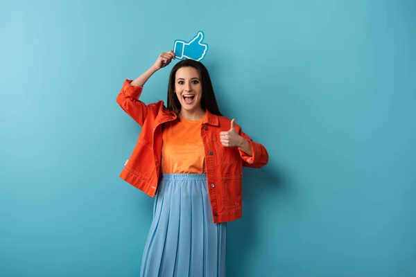 Smiling woman showing thumb up and holding paper like on blue background — Stock Photo