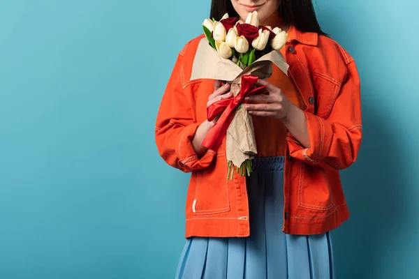 Cropped view of smiling woman in jacket smelling bouquet on blue background — Stock Photo