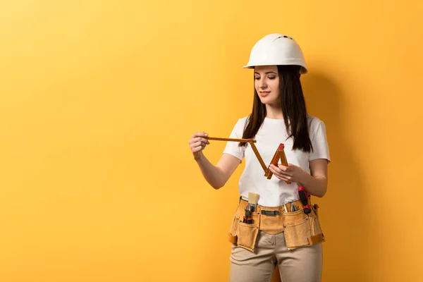 Lächelnde Handarbeiterin im Helm mit Messgerät auf gelbem Hintergrund — Stockfoto