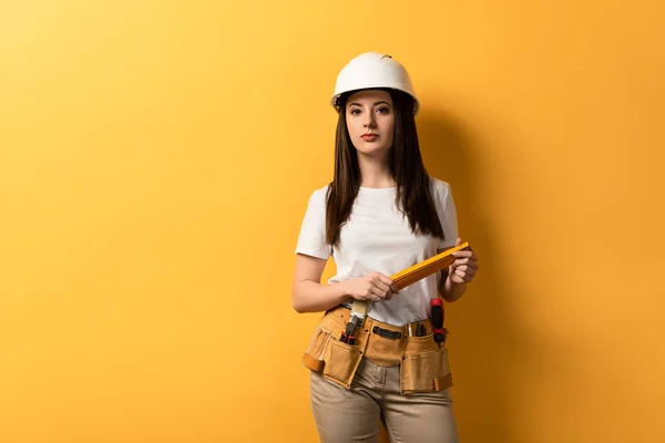 Handarbeiterin im Helm hält Messgerät in der Hand und blickt auf Kamera auf gelbem Hintergrund — Stockfoto