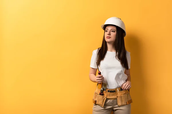 Atraente handywoman segurando chave de fenda e olhando para longe no fundo amarelo — Fotografia de Stock