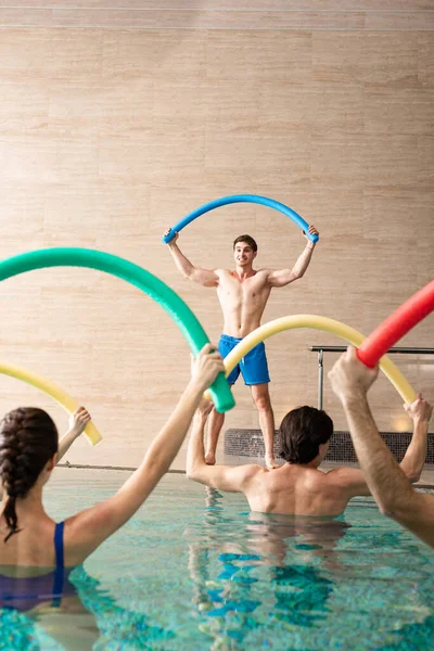 Treinador sorridente com macarrão de piscina exercitando com grupo de jovens na piscina — Fotografia de Stock