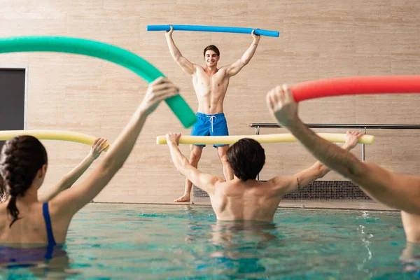 Selective focus of handsome trainer smiling while training group of young people with pool noodles in swimming pool — Stock Photo