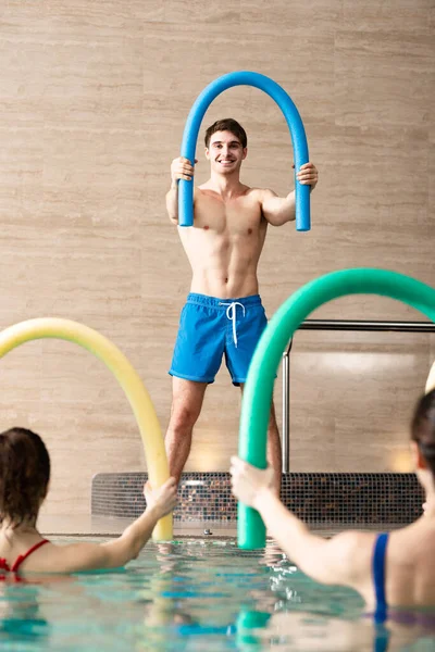 Selective focus of smiling trainer exercising with pool noodle and group of people in swimming pool — Stock Photo