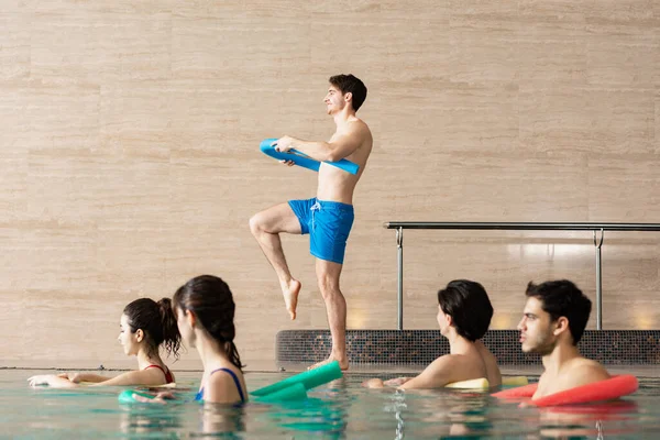 Side view of trainer with pool noodle working out with group of people during water aerobics in swimming pool — Stock Photo