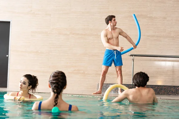 Enfoque selectivo del entrenador sosteniendo fideos de la piscina mientras hace ejercicio con personas en la piscina - foto de stock