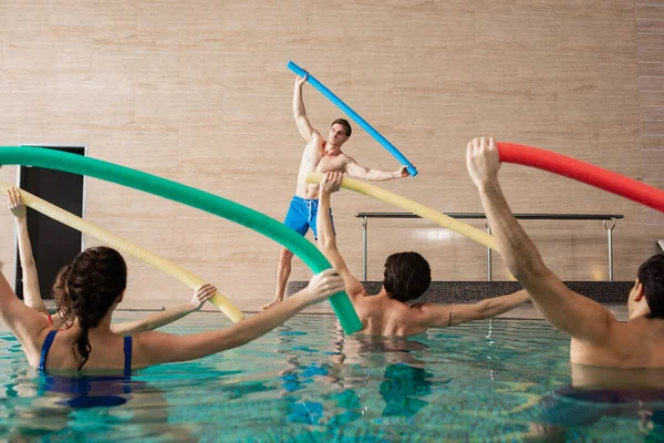 Selective focus of group of people holding pool noodles while exercising with trainer in swimming pool — Stock Photo