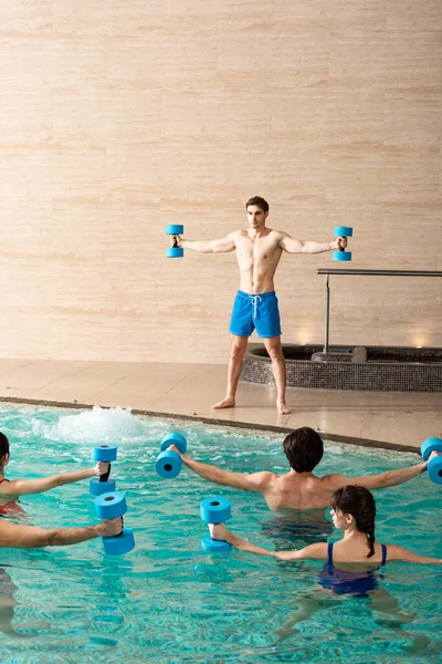 Guapo entrenador sosteniendo mancuernas durante aeróbic acuático con grupo de personas en la piscina - foto de stock