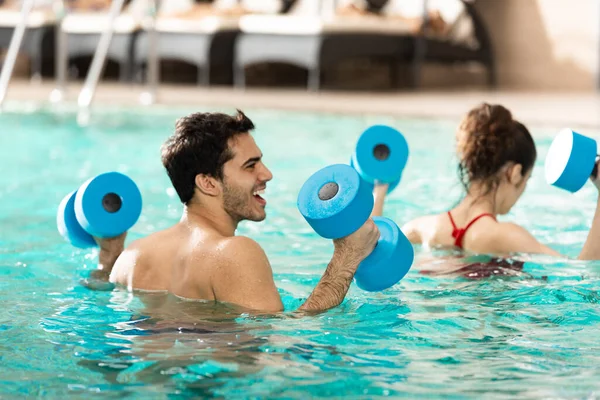 Selective focus of cheerful man with dumbbells during water aerobics in swimming pool — Stock Photo