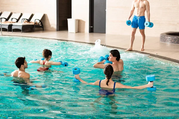 Groupe de jeunes avec haltères faisant de l'aquagym avec entraîneur dans la piscine — Photo de stock