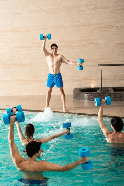 Allenatore sorridente che mostra esercizio con manubri a gruppo di persone in piscina — Foto stock