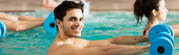 Vista lateral del hombre sonriendo a la cámara mientras hace ejercicio con pesas en la piscina, plano panorámico — Stock Photo