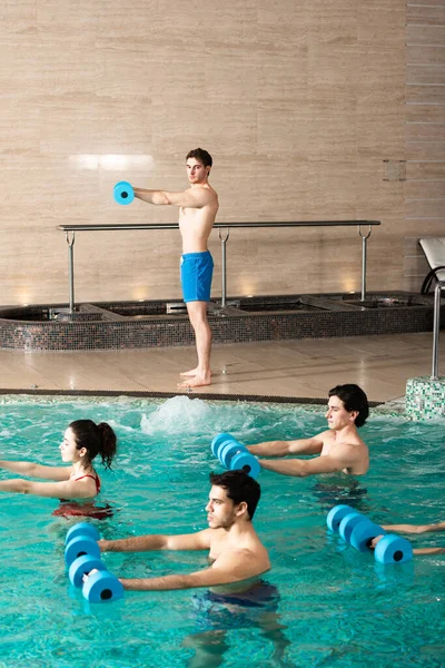 Vista lateral do treinador segurando halteres durante o exercício com grupo de jovens na piscina — Fotografia de Stock