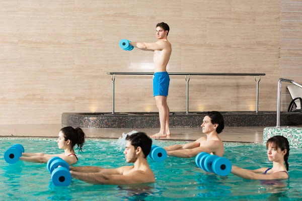 Seitenansicht einer Gruppe junger Leute, die mit Kurzhanteln im Schwimmbad in der Nähe des Trainers trainieren — Stockfoto