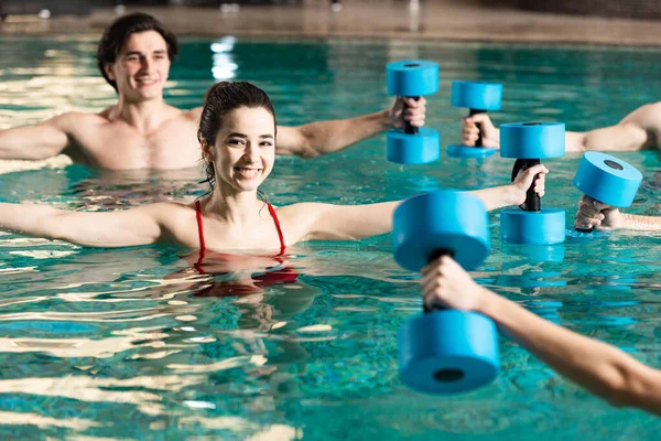 Selective focus of smiling young people exercising with barbells during water aerobics in swimming pool — Stock Photo
