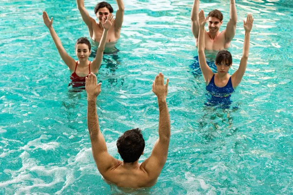 Overhead view of trainer with raised hands showing exercise to smiling young people in swimming pool — Stock Photo