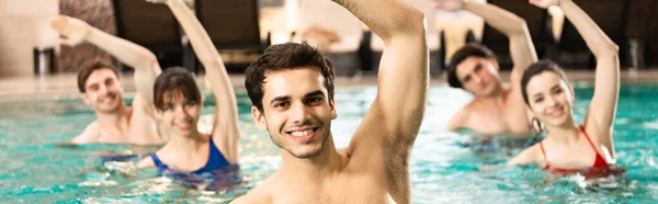 Selective focus of handsome trainer smiling at camera while exercising with people in swimming pool, panoramic shot — Stock Photo