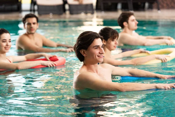 Side view of smiling people with pool noodles training in swimming pool — Stock Photo