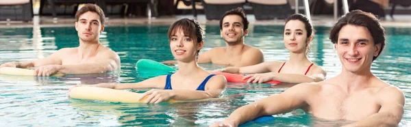 Panoramic shot of group of young people smiling at camera while training with pool noodles in swimming pool — Stock Photo