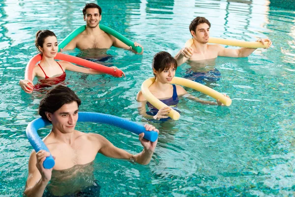 Sonrientes jóvenes haciendo ejercicio con fideos de la piscina durante aeróbicos acuáticos en la piscina - foto de stock