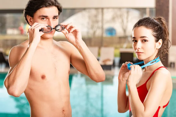 Man and woman looking at camera while holding swimming googles near swimming pool — Stock Photo