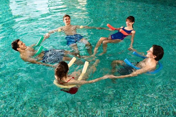 Blick aus der Vogelperspektive auf lächelnde junge Leute, die Händchen halten, während sie im Schwimmbad mit Poolnudeln trainieren — Stockfoto