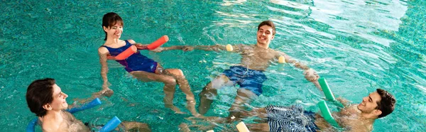 Foto panorámica de jóvenes sonrientes tomados de la mano mientras hacen ejercicio con fideos de piscina en la piscina - foto de stock