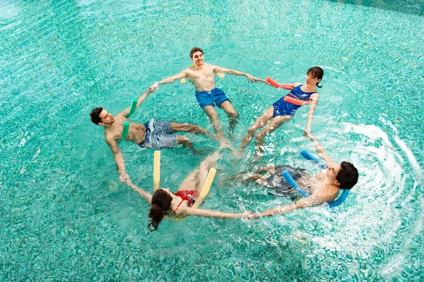 Blick aus dem Hochwinkel auf eine Gruppe junger Leute, die Händchen halten, während sie Wassergymnastik mit Poolnudeln im Schwimmbad trainieren — Stockfoto