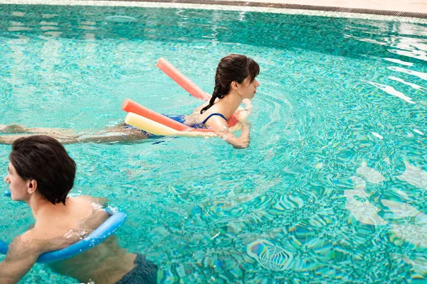 Side view of man and woman swimming in pool with pool noodles — Stock Photo