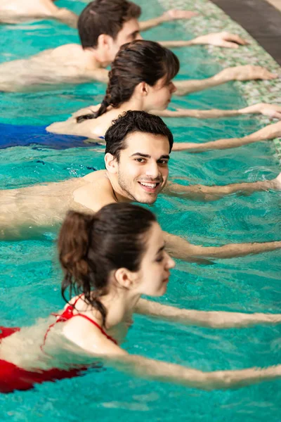 Вид сбоку красивого человека, улыбающегося на камеру во время тренировки с молодыми людьми в бассейне — стоковое фото
