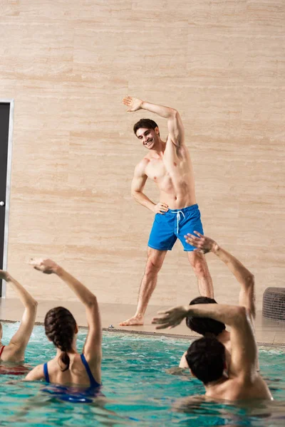 Вибірковий фокус усміхненого тренера, який дивиться на групу молодих людей під час водної аеробіки в басейні — стокове фото