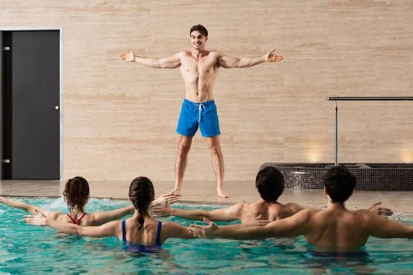 Entrenador sonriente mostrando ejercicio a grupo de jóvenes en piscina - foto de stock