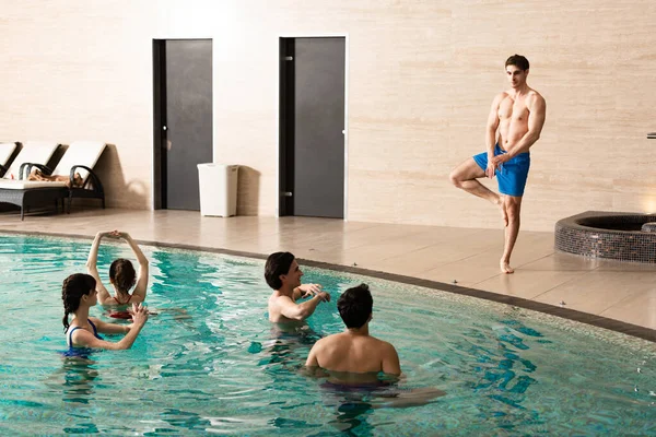 Handsome trainer standing on one leg while showing exercise to young people in swimming pool — Stock Photo