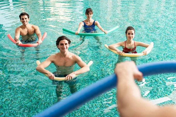 Foco seletivo de pessoas sorridentes com macarrão de piscina olhando para o treinador durante a aeróbica aquática na piscina — Fotografia de Stock