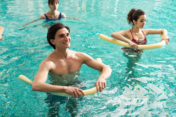 Foco seletivo de homem sorridente segurando macarrão da piscina enquanto treina com mulheres na piscina — Fotografia de Stock