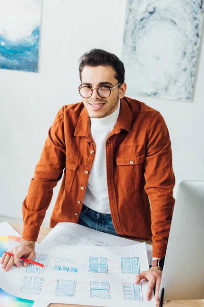 Schöner UX-Designer lächelt in die Kamera, während er am Tisch mit Webseiten-Vorlagen arbeitet — Stockfoto