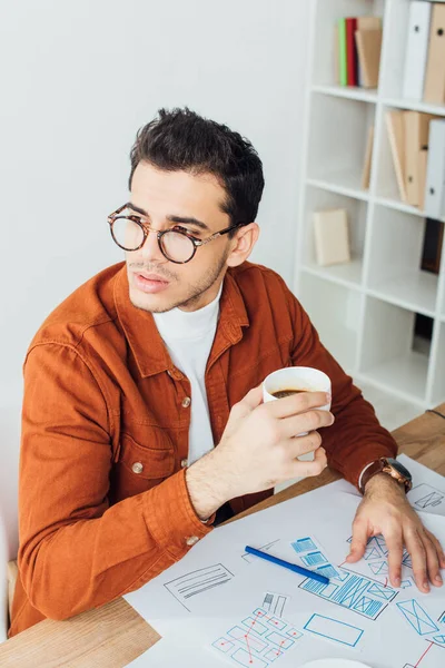 Дизайнер п'є каву біля ескізів дизайну користувача на столі — стокове фото