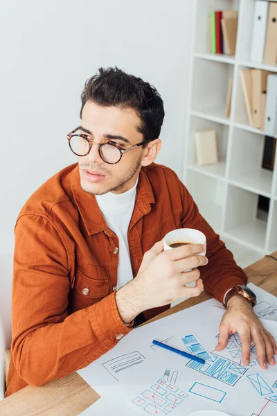 Diseñador beber café cerca de bocetos de diseño de experiencia de usuario en la mesa - foto de stock