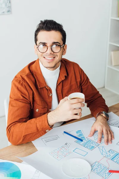 Designer holing tasse à café et souriant à la caméra près des modèles de ux projet sur la table — Photo de stock