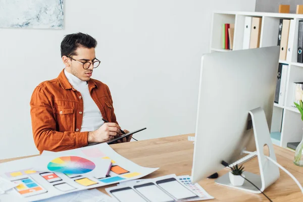 Handsome creative designer using graphics tablet near computer and layouts of user experience design on table — Stock Photo